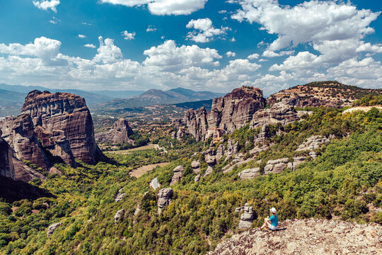 View of towering rock formations piercing the skyline, lush greenery blanketing the terrain, a lone figure gazing at the landscape, Kalabaka, Trikala, Greece.