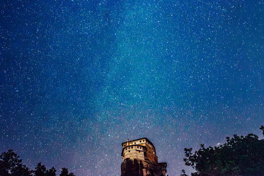 View of the illuminated monastery perched atop a rugged, rocky pinnacle under a vast, twinkling expanse of the night sky, Kalabaka, Trikala, Greece.