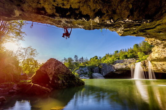 View of a daring climber suspended upside down from a cave mouth above a serene green lake, framed by lush trees and a cascading waterfall, Pazin, Istria County, Croatia.