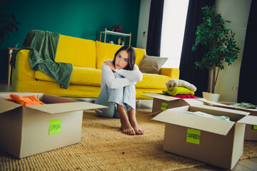 Young woman sits on the floor surrounded by moving boxes in a bright living room with couch tired...