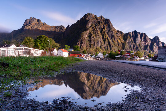 View of jagged peaks reflected in a roadside puddle after a rain, with the vibrant hues of houses nestled at the base, Lofoten Islands, Nordland, Norway.