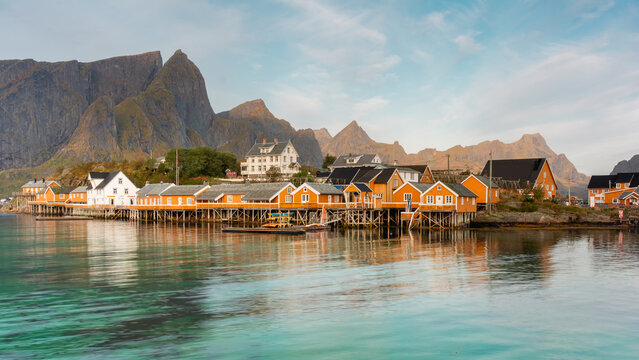 View of vibrant yellow cabins on stilts mirroring in the tranquil turquoise water, against the backdrop of jagged peaks, Lofoten Islands, Nordland, Norway.