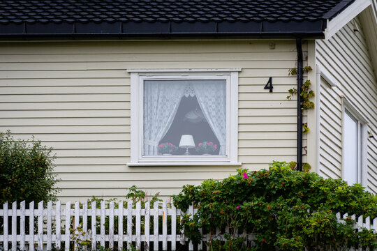 View of a cozy window adorned with lace curtains, a lamp, and flowers, nestled in a quaint house with a white picket fence, Lofoten Islands, Nordland, Norway.