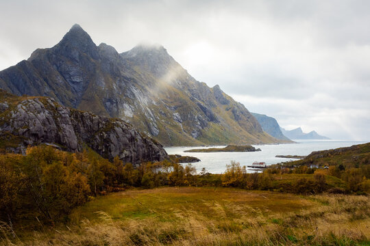 View of jagged peaks rise majestically against a cloudy sky, the sun's rays piercing through, illuminating the autumn foliage along the serene coastline, Lofoten Islands, Norway.