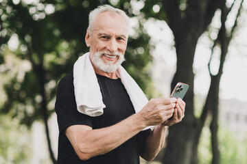 Fototapeta premium Elderly bearded man athletic shirt smiles while checking his smartphone in sunlit park after workout towel shoulders enjoying outdoor fitness fresh air