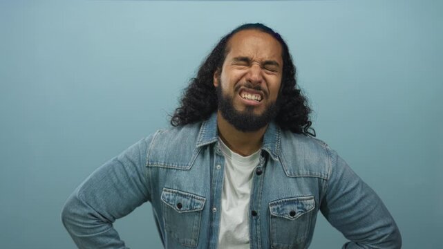 Man wearing denim jacket with hands on hips looking down in a pale blue studio; frustration fatigue.