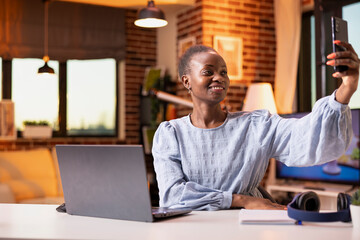 Obraz premium Cheerful female employee working remotely takes selfie with smartphone during break at home office desk. Smiling female entrepreneur raises mobile device to capture photo for social media.