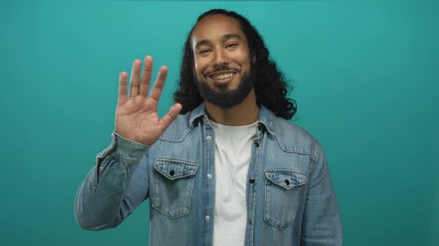 Hispanic arab man wearing denim jacket waving raised hand at camera in teal studio with visible palm; friendly greeting.