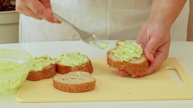Female hands making sandwich with mashed avocado slices and wholegrain bread for breakfast
