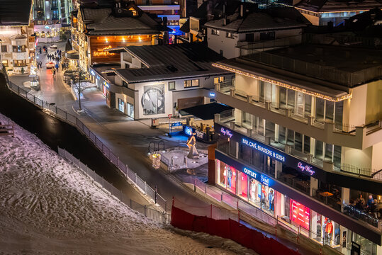 Snow lined pedestrian Ischgl promenade in a European alpine resort at night, with neon and fairy lights, modern bar cafe windows, chalet facades, and adjacent ski run.
