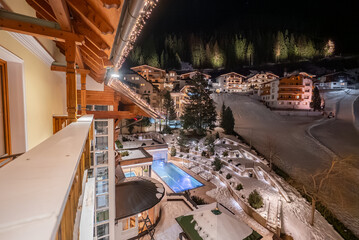 Fototapeta premium Nighttime view from a timber balcony shows an alpine hotel complex in Ischgl with a heated blue pool, snow terraces, pine trees, lit paths, steam from a spa, and a ski slope in winter.