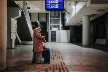A solo traveler stands with a blue suitcase looking up at the departures board. The lone passenger waits in an empty station concourse beneath overhead signage. © qunica.com