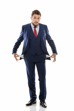 Full length portrait of a worried young businessman in a navy blue suit showing empty pockets, bankrupt man with no money standing isolated on white background.