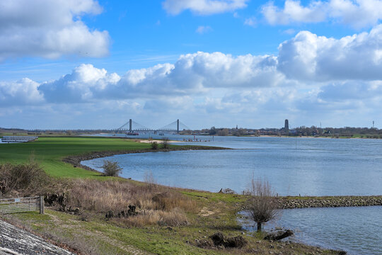 Panoramic view of the Waal river in the Netherlands with stone groynes, green floodplains and a modern cable-stayed bridge under a dramatic cloudy sky, Dutch river landscape