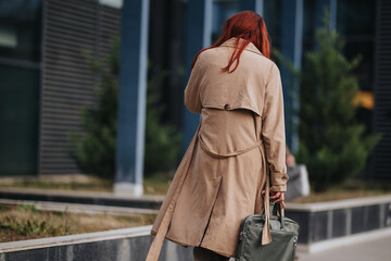 Naklejka premium A redheaded woman in a trench coat walks away carrying a briefcase toward an urban office. The professional commuter heads to a business meeting in a modern city setting.