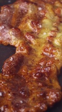 Slices of bacon frying in black pan closeup, vertical video format