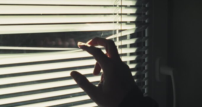 Tight, dramatic shot of fingers parting Venetian blinds to peer outside from a dark interior. Evokes feelings of fear, invasion, and classic stalker thriller tropes.