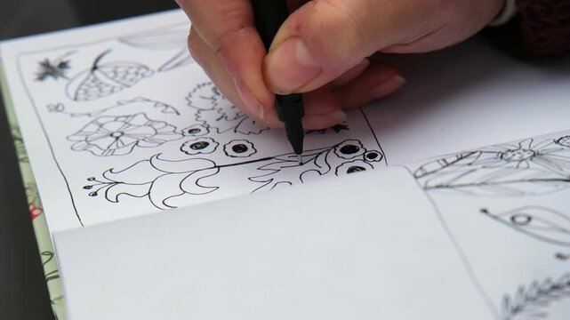 Close-up of a hand carefully sketching detailed flower patterns and leaves in a sketchbook. The movement is steady as the pen creates thin, precise black lines on a white page.