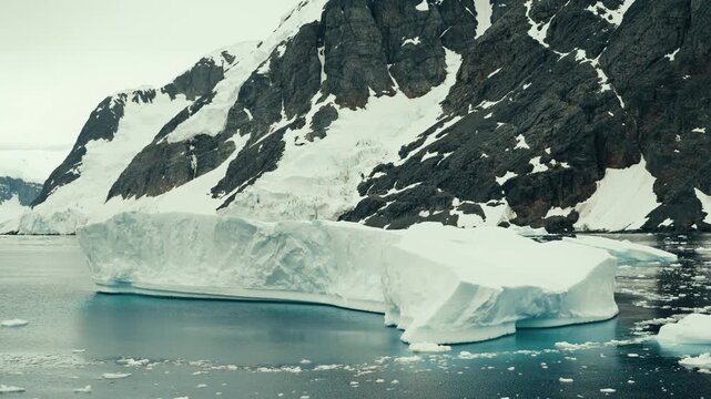 Massive tabular iceberg drifts through glassy teal waters of Lemaire Channel with dramatic snow-covered rocky cliffs rising behind in slow cinematic parallax shot