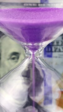 Purple sand dropping through the bulb of the hourglass and dollar money on the background. Hourglass with sand stand among money. Deadline, business planning, finance and time is money concept.
