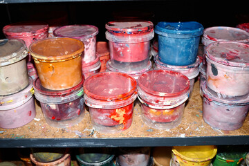 Stacked plastic containers filled with various colors of thick paint sitting on wooden shelf,...