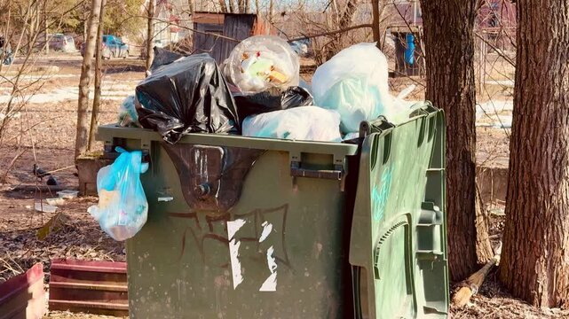Overflowing green dumpster on sidewalk, piled with blue plastic bags and black sacks, rust streaked metal container marked.