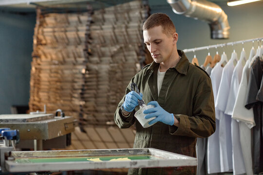 Caucasian young adult man preparing screen printing ink while standing in workshop, wearing protective gloves, focusing on mixing process with clothing items hanging in background