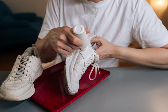 Closeup of shoemaker using bottle of shoe whitener and painting scuffed surface of dirty white sneaker, illustrating process of cleaning, recoloring, and restoring footwear