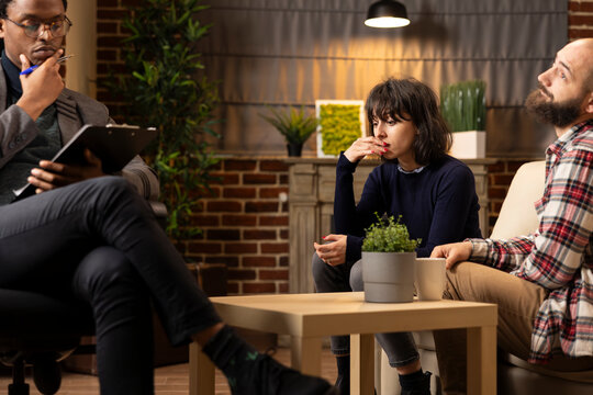 Worried woman with hand on mouth sits on couch during therapy session as African American therapist reads notes and boyfriend listens. Couple receives relationship assessment from psychiatrist indoors