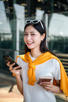 Young asian woman traveler using smartphone for checking flight information check the routes trip while waiting with her luggage at the airport terminal. Traveller woman passport and baggage luggage