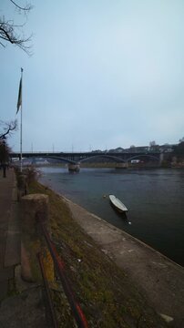 Vertical static shot of a traditional wooden boat anchored on the Rhine river with the Wettsteinbruecke bridge in the background on a foggy morning.
