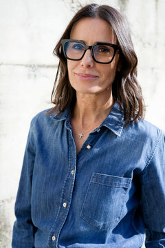 Woman with glasses wearing denim shirt standing against a light textured wall. Milan, Italy