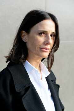 Woman in black coat and white shirt stands against a light gray background, looking confident. Milan, Italy
