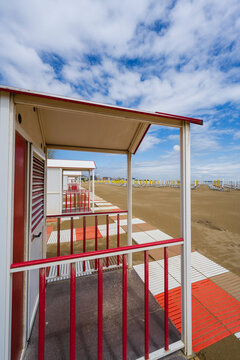Red and white beach huts line a sandy shoreline under a vibrant blue, cloudy sky. Caorle, Metropolitan City of Venice, Veneto, Italy