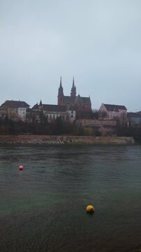 Vertical static shot of the Basel Minster (Basler M&uuml;nster) cathedral on a hill above the Rhine river with colorful buoys in the foreground on a misty day.