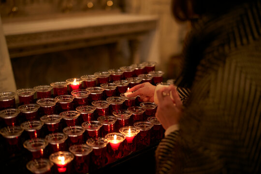 Rows of lit candles in red glass holders with person lighting one in a dimly lit room. Zurich, Switzerland