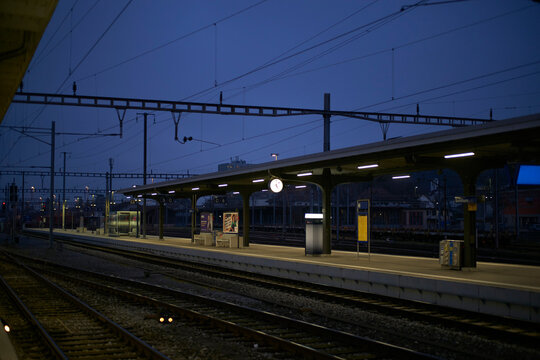 Empty railway station platform at dusk with overhead wires and a clock under a deep blue sky. Zurich, Switzerland