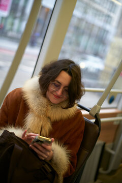 Woman in fur-lined coat using a smartphone on public transport, smiling and looking down. Brussels, Belgium