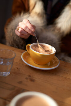 Hand stirring a hot beverage in a yellow cup on a wooden table with fur coat visible. Brussels, Belgium