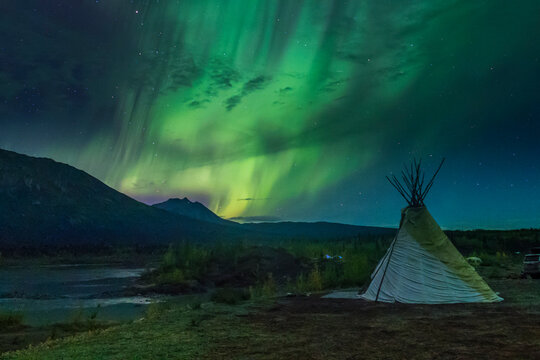 Teepee under vibrant green aurora in a mountainous wilderness landscape at night. Wrangell St. Elias National Park, Alaska, USA
