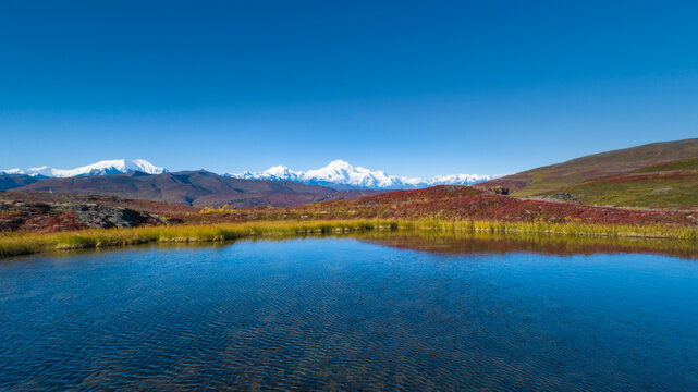 A serene mountain landscape with a clear lake, vibrant fields, and snow-capped peaks under a blue sky. Wrangell St. Elias National Park, Alaska, USA