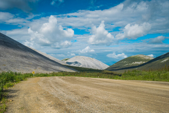 Curving gravel road through a mountainous landscape under a vivid blue sky with fluffy clouds. Canada