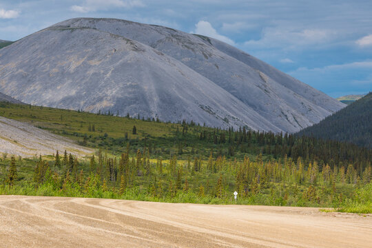 Expansive view of a mountain landscape with a gravel road and forested area under a cloudy sky.  Yukon Territory, Canada