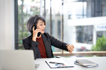 Obraz premium Smiling asian woman checking time while making phone call , confident businesswoman planning meeting with partner, looking at wristwatch, task management.