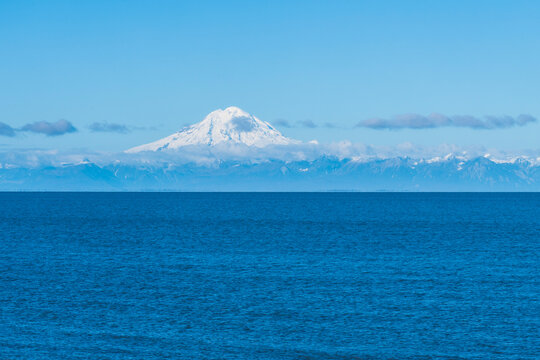 Snow-covered mountain peak over a calm blue ocean with clear sky and distant clouds. Anchor River, Alaska, USA