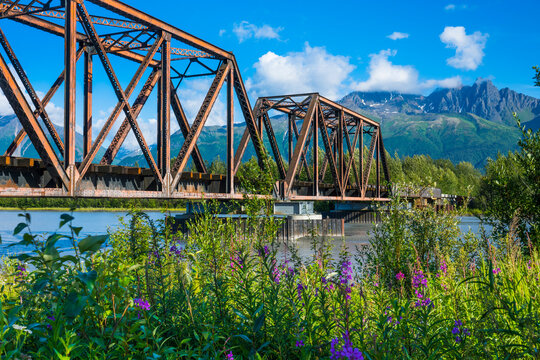 A rusted metal bridge spans a river with a backdrop of mountains under a clear blue sky. Palmer, Alaska, USA