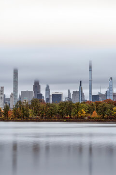 City skyline with tall skyscrapers behind a row of autumn trees reflected on calm water. Central Park Lake, New York CIty, USA