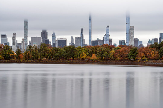 City skyline with tall buildings obscured by clouds above a colorful autumn forest and calm water. Central Park Lake, New York CIty, USA