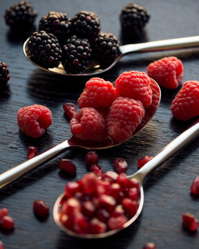 Close-up of spoons with raspberries, blackberries, and pomegranate seeds on a wooden surface.