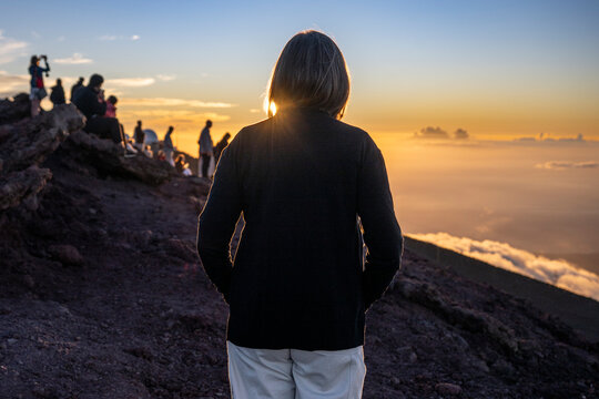 Woman stands on rocky ground at sunrise, with people watching the horizon. Kula, Maui, Hawaii